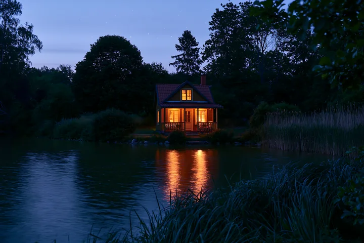A wooden holiday home under stars lights up from within as it sits by a calm lake.