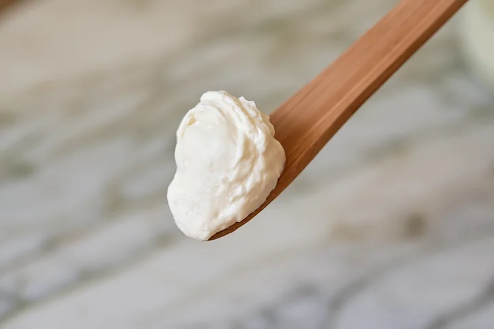 A wooden spoon holds a cream dose with matte texture, placed on a light-colored marble surface.