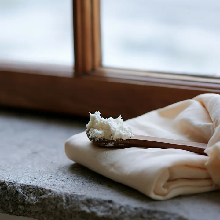 A wooden spoon with cream is placed beside cream-colored fabric on a stone surface near a window.