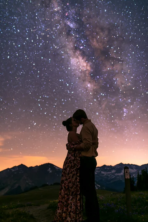 A young couple kissing beneath visible Milky Way in a clear night sky.