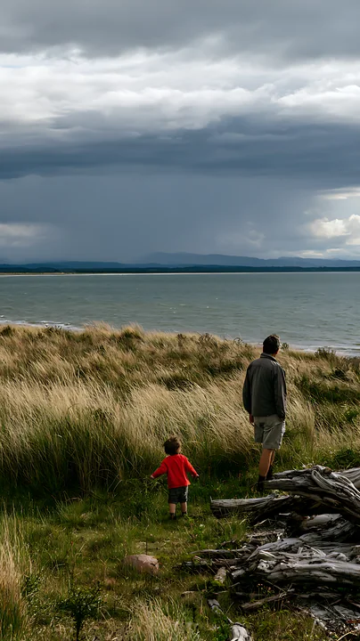 Adult and child walk through coastal grass toward the sea beneath dramatic storm clouds.