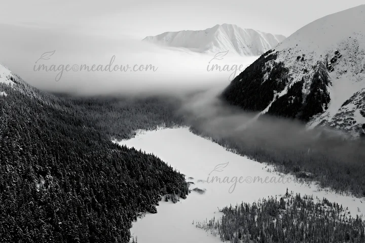 An aerial view of a snowy meadow valley, surrounded by mountains, with mist hovering above the landscape, adding depth and dimension to the view. Presented as a