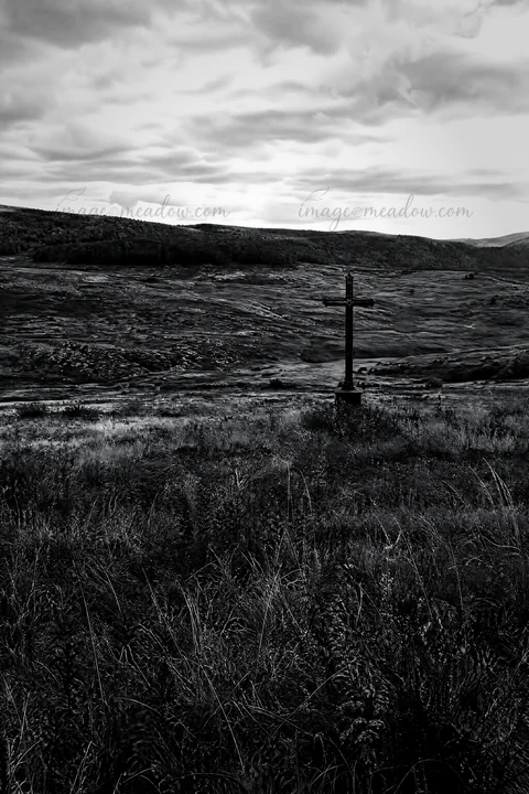 An aged Christian cross stands in a meadow located in a deep mountain valley during autumn. The landscape features hills and patches of grass under a cloudy sky