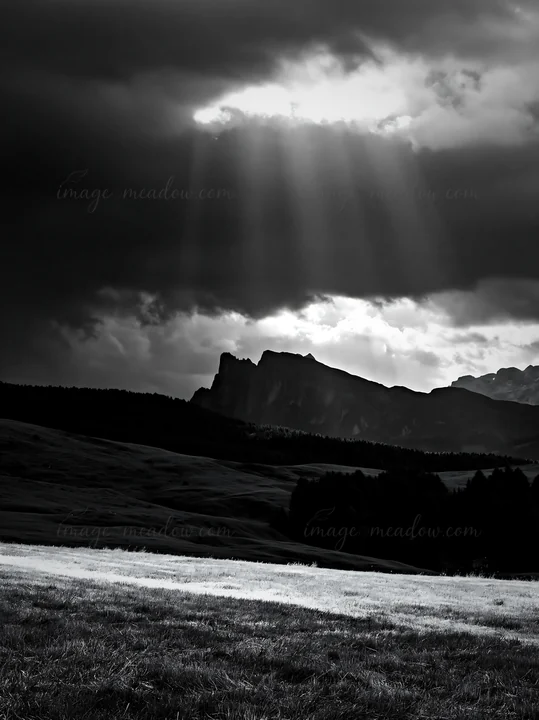 Sunbeams break through dark clouds over the Alpe di Siusi meadow, illuminating the green landscape and nearby mountains. The scene captures a moment of changing