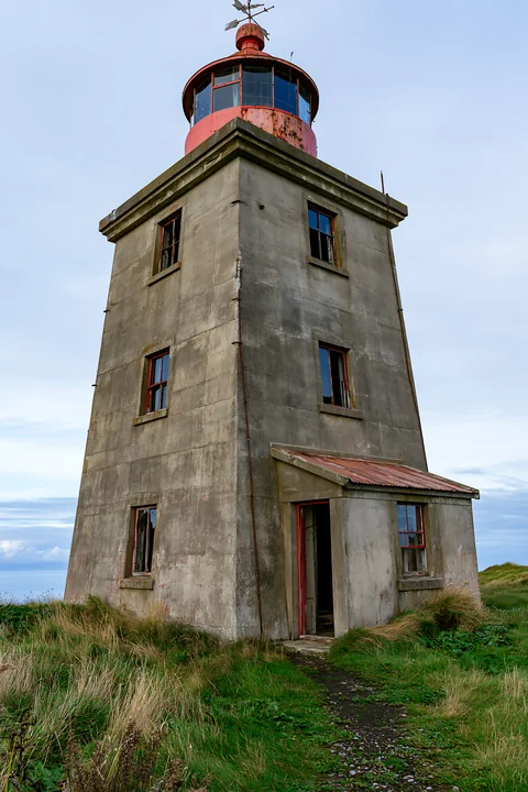An abandoned lighthouse on a remote coast stands tall and weathered, surrounded by grass and a cloudy sky.