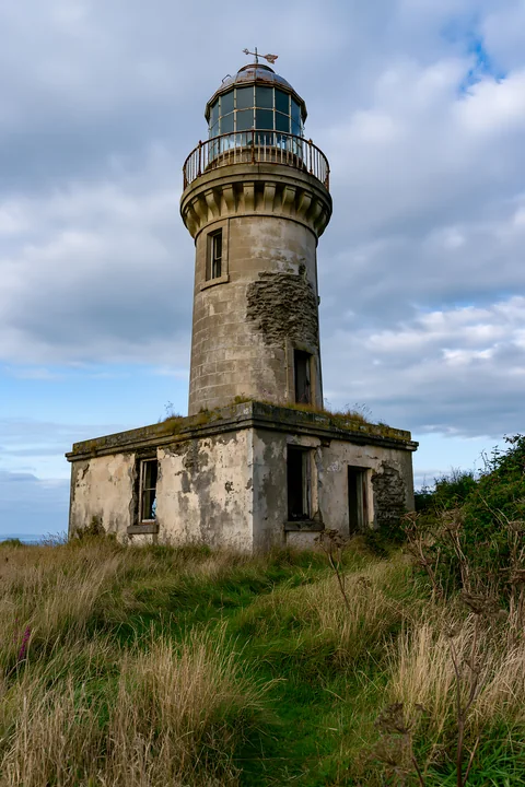 An abandoned lighthouse on a remote coast stands tall with patches of grass surrounding its base.