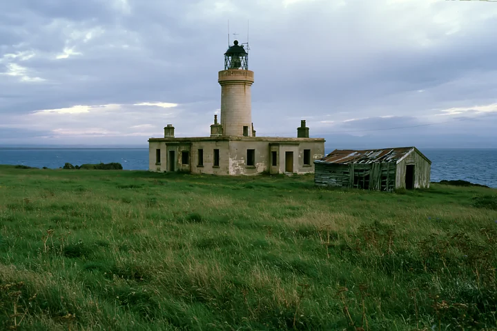 An abandoned lighthouse stands on a remote coast, with an old shed nearby.