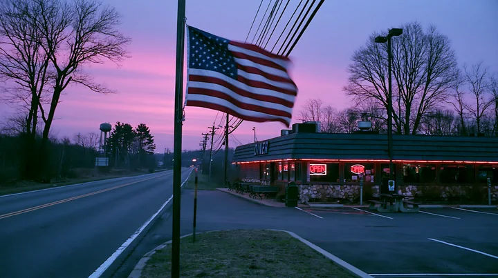 An American flag flying in strong wind is visible next to a diner along a road, with a colorful sunset in the background.