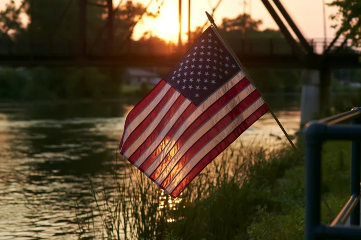 An American flag flying in strong wind stands near the water as the sun sets behind trees and a bridge.
