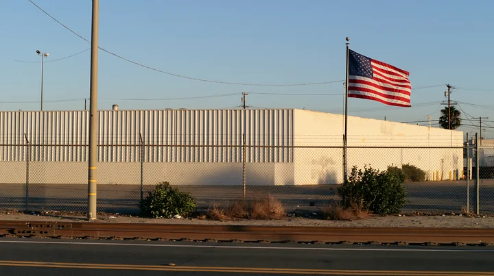 An American flag outside a factory is seen near a fence and road.