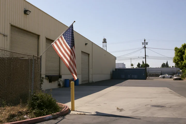 An American flag waves on a pole next to a factory in an industrial area.