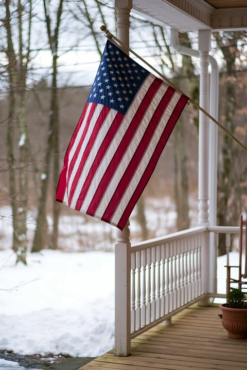 An American flag waving in gentle breeze on a porch surrounded by snowy trees.
