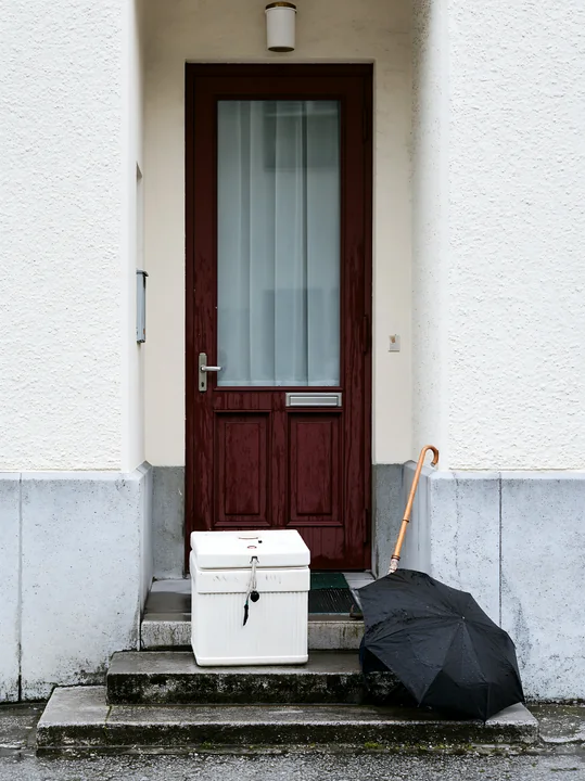 An apartment doorstep shows an insulated milk delivery box placed next to a black umbrella.