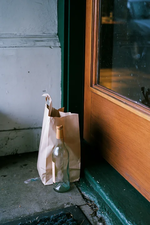 An empty bottle after party sits beside a brown paper bag near a closed wooden door.