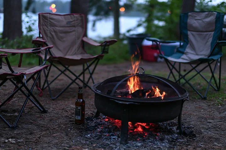 An empty bottle sits beside a campfire in a wooded area.