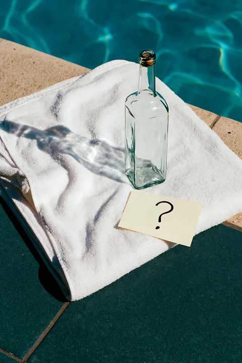 An empty bottle with question mark note rests on a towel beside a pool.