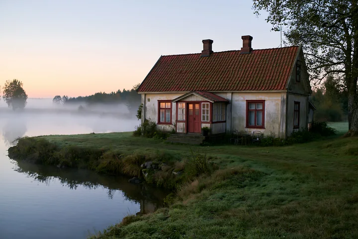 An old cottage with weathered facade stands by a quiet waterway at dawn.