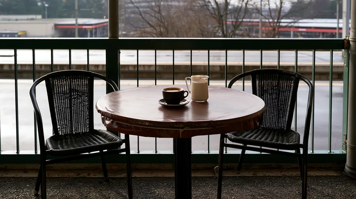 An outdoor cafe table holds a latte and a milk jug with two black chairs nearby.