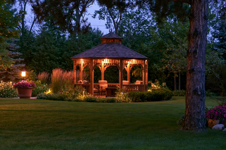 An upscale garden pavilion provides a cozy seating area surrounded by a well-kept lawn and lush greenery at dusk.