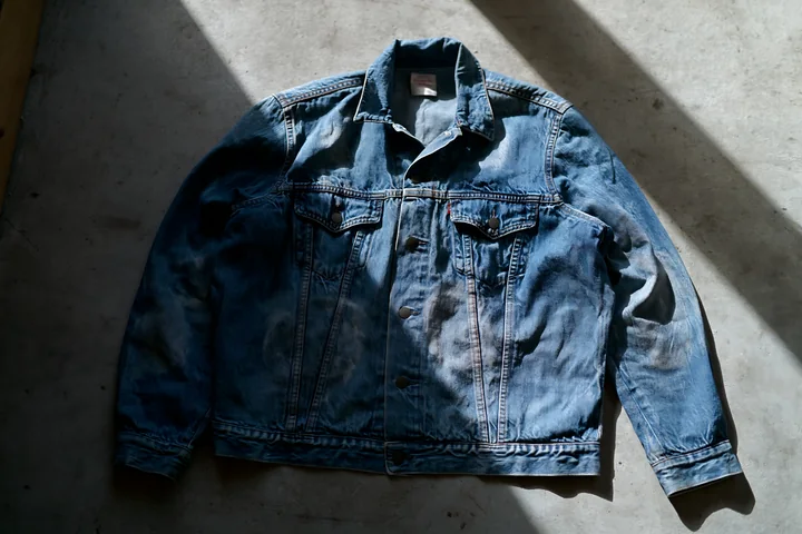 Blue denim jacket with dried watermarks lies on a concrete floor in strong diagonal sunlight.