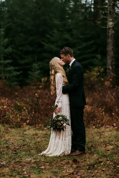 Bride and groom kiss in wedding attire while standing with a bouquet in a leafy forest.