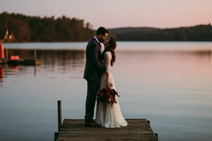 Bride and groom kiss on a wooden dock beside a calm lake at sunset.