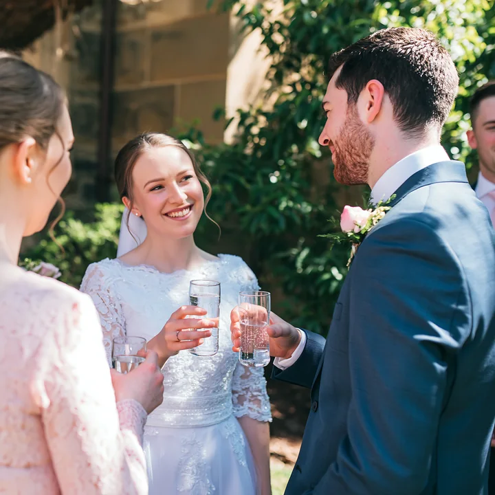 Bride, groom, and guests raise glasses together outdoors in bright light near a church.