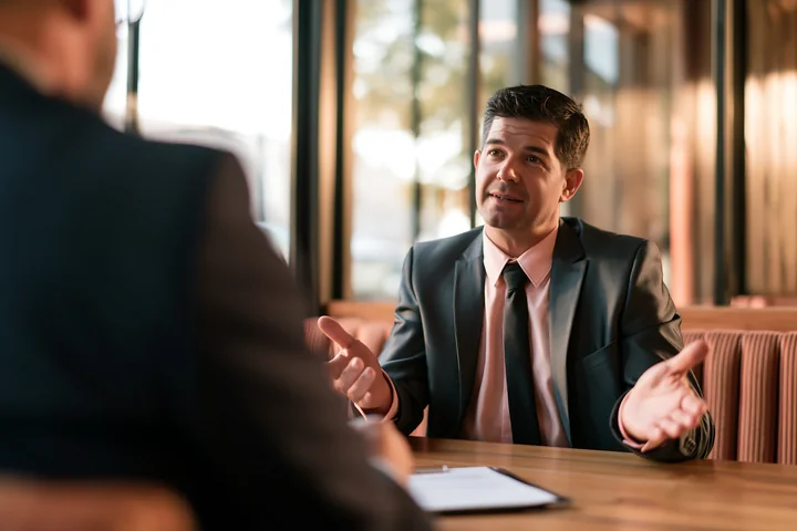 Businessman discusses paperwork with an advisor across a table in a bright professional meeting space.