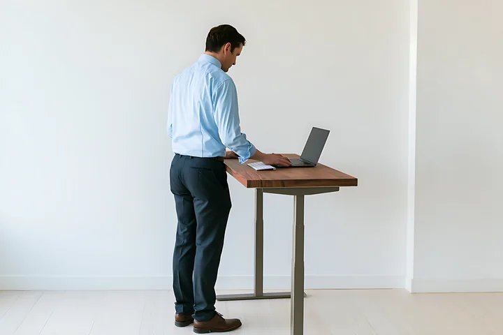 Businessman stands alone at a wooden desk using a laptop in a bright minimalist office.