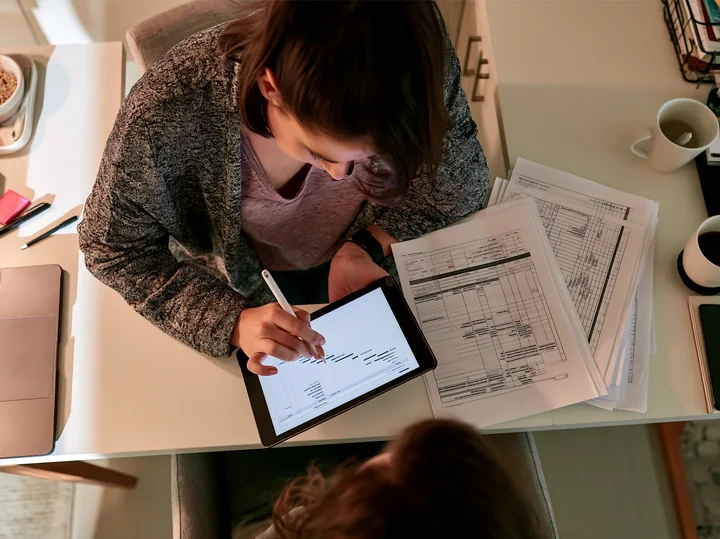 Businesswoman reviews printed schedules and marks a tablet with a stylus at a desk.