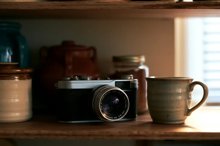 Camera and lens on a wooden shelf sit alongside a ceramic cup and various jars.