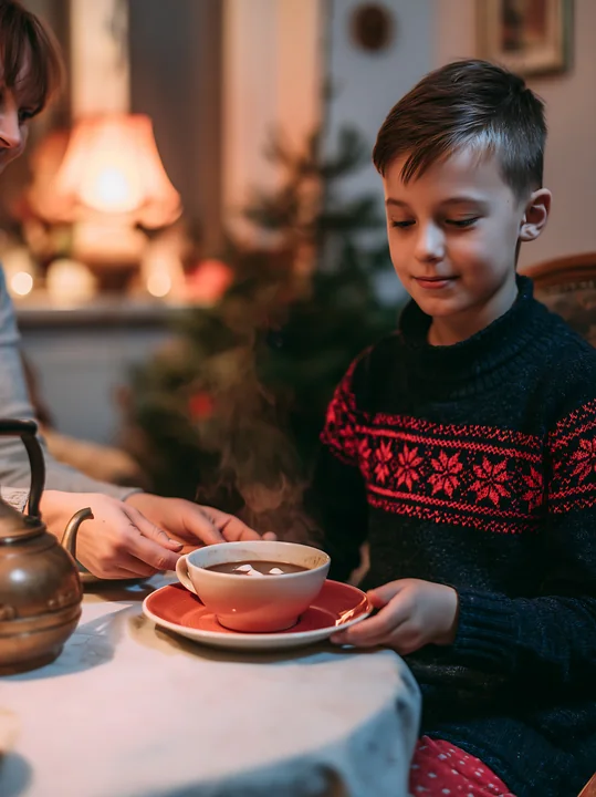 Child sits beside a Christmas tree while an adult serves steaming cocoa with marshmallows at a table.