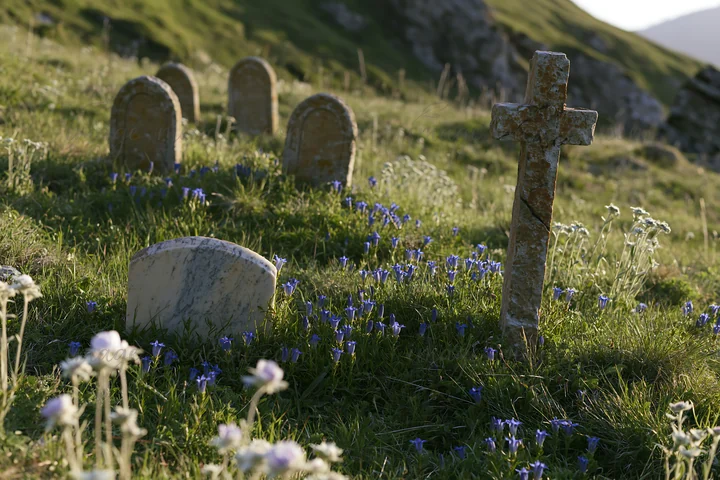 A Christian cross stands in a sunlit alpine meadow surrounded by tombstones and small wildflowers. The scene captures the natural landscape and religious symbol