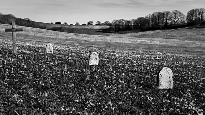 A Christian cross stands in a winter meadow surrounded by snowdrops. The sun sets low on the horizon, casting light rays across the landscape with rolling hills