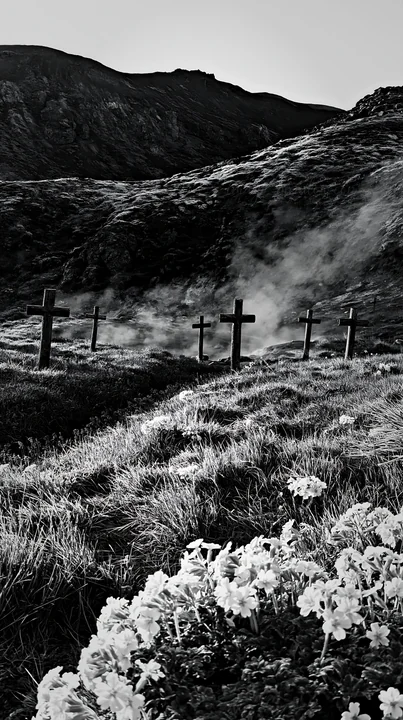 Christian crosses stand in a meadow near a geothermal spring, surrounded by green grass and flowers. The setting features smoke rising from the spring, with mou
