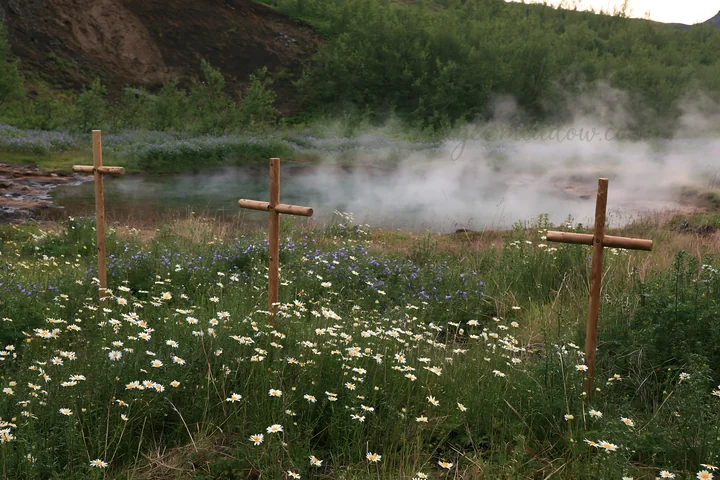 Three wooden crosses stand in a meadow near a geothermal spring. Steam rises from the spring, surrounded by wildflowers in bloom. This location is characterized