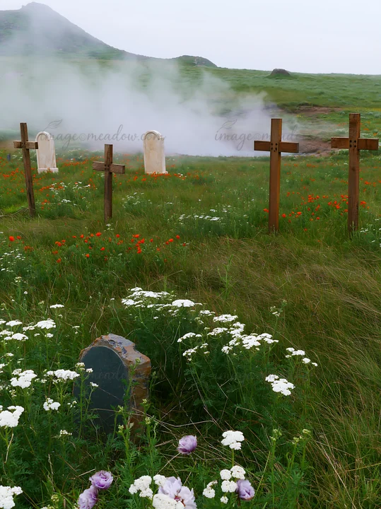 Wooden Christian crosses stand in a meadow near a geothermal spring. Flowers surround the area, adding color to the landscape. A hill is visible in the backgrou