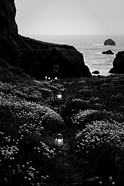 A pathway lined with lanterns leads to a cave in a meadow close to the ocean. The scene is dusk with low light, highlighting the soft glow of the lights among t