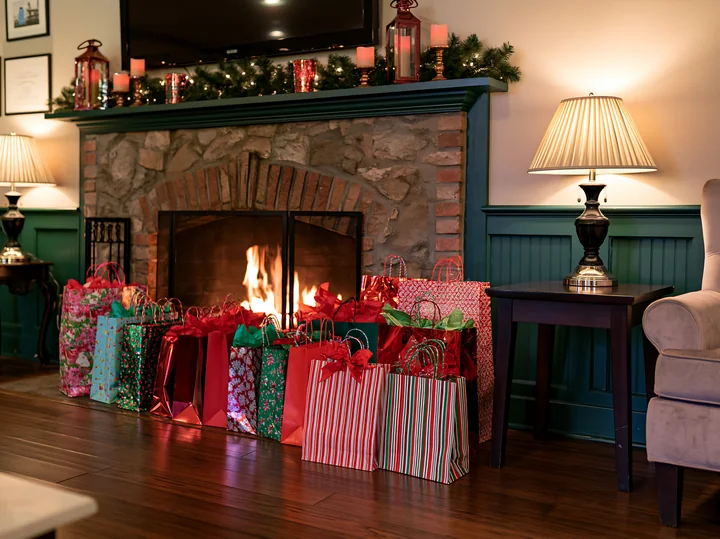 Colorful holiday shopping bags sit on the floor beside a lit stone fireplace in a warmly decorated room.