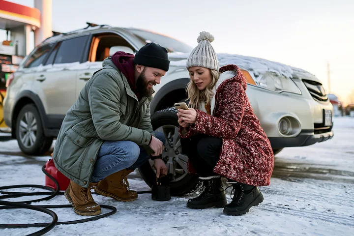 Couple checks a smartphone beside a snow-covered car during winter road trip preparation.