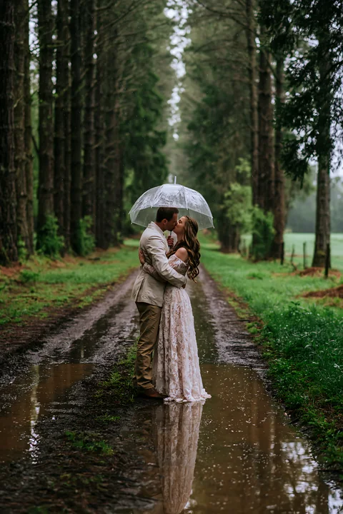 Couple kisses beneath a clear umbrella on a muddy forest road during rain.