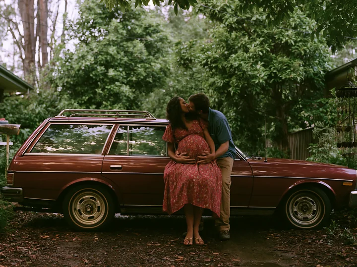Couple kisses beside a vintage station wagon in a leafy wooded setting.