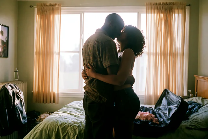 Couple kissing and embracing beside an unmade bed in a bright bedroom with sunlight through the window.