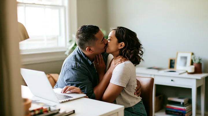 Couple shares a tender kiss beside an open laptop in a bright home office.