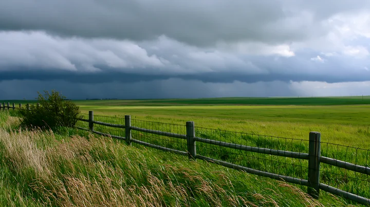 Dark storm clouds gather over a green meadow, with a wooden fence in the foreground.