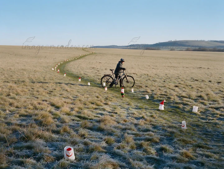 A cyclist rides through a meadow during daylight. The path is marked by colorful Christmas lanterns. The scene shows the frost-covered grass and distant hills.