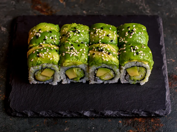 Dragon roll close-up features sushi rolls with avocado and cucumber, garnished with sesame seeds, served on a dark stone plate, ideal for food photography and s