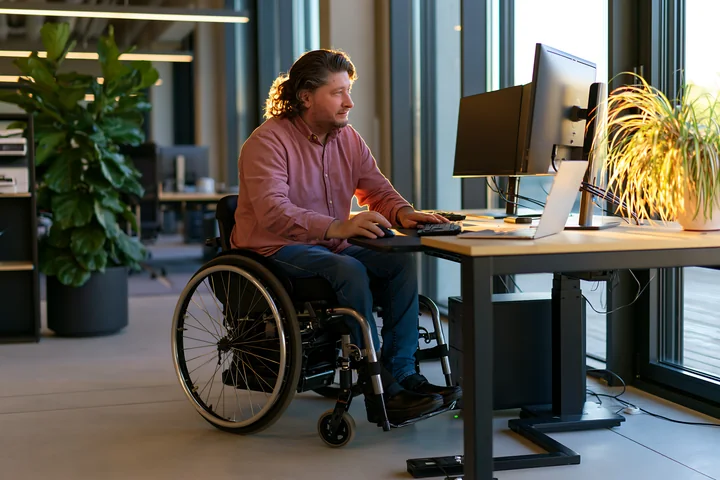Employee using a wheelchair works at a computer workstation in a bright, plant-filled modern office.