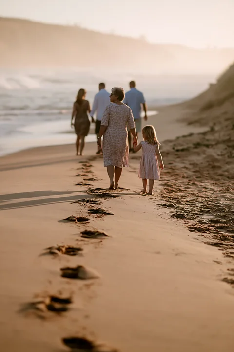 Family members walk along a sandy beach at sunset beside the ocean, leaving footprints in the wet sand.