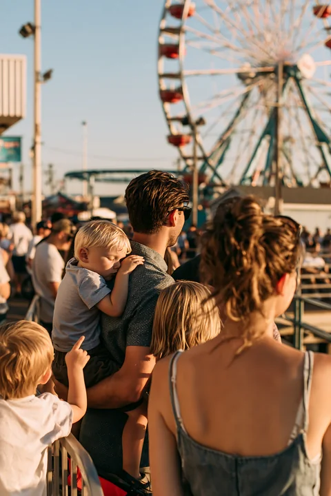 Family waits in an amusement park queue while a child is held near a ferris wheel.